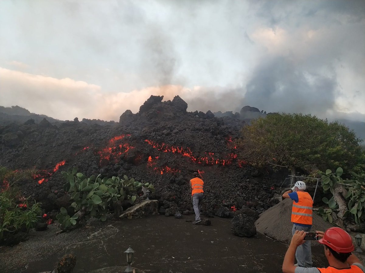 Two people in high visibility clothing stand next to a glowing lava flow, which is about twice their height 