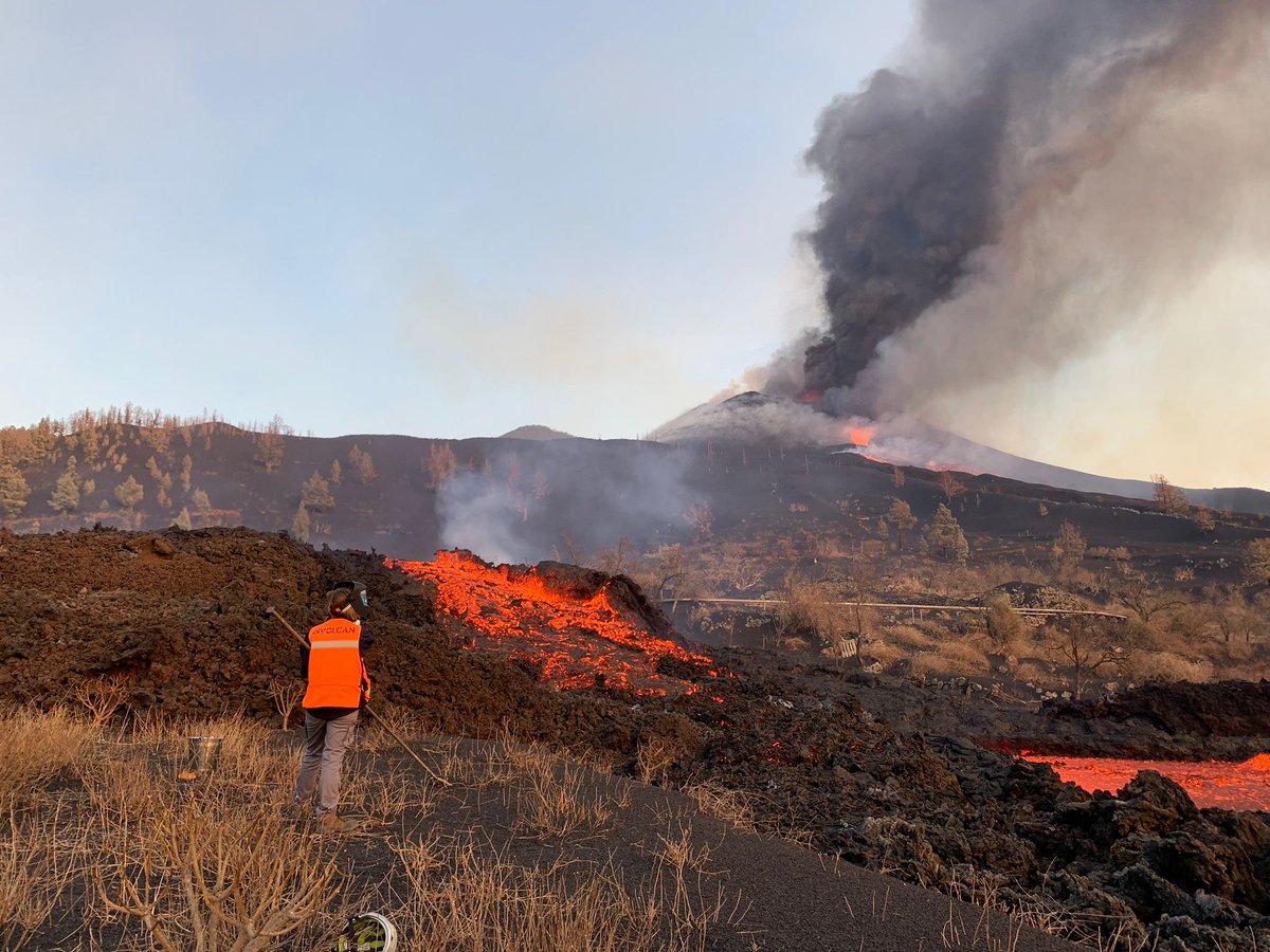 A scientist stands next to a large, orange, fast flowing lava flows. The erupting volcano and its dark grey ash cloud is seen in the background.
