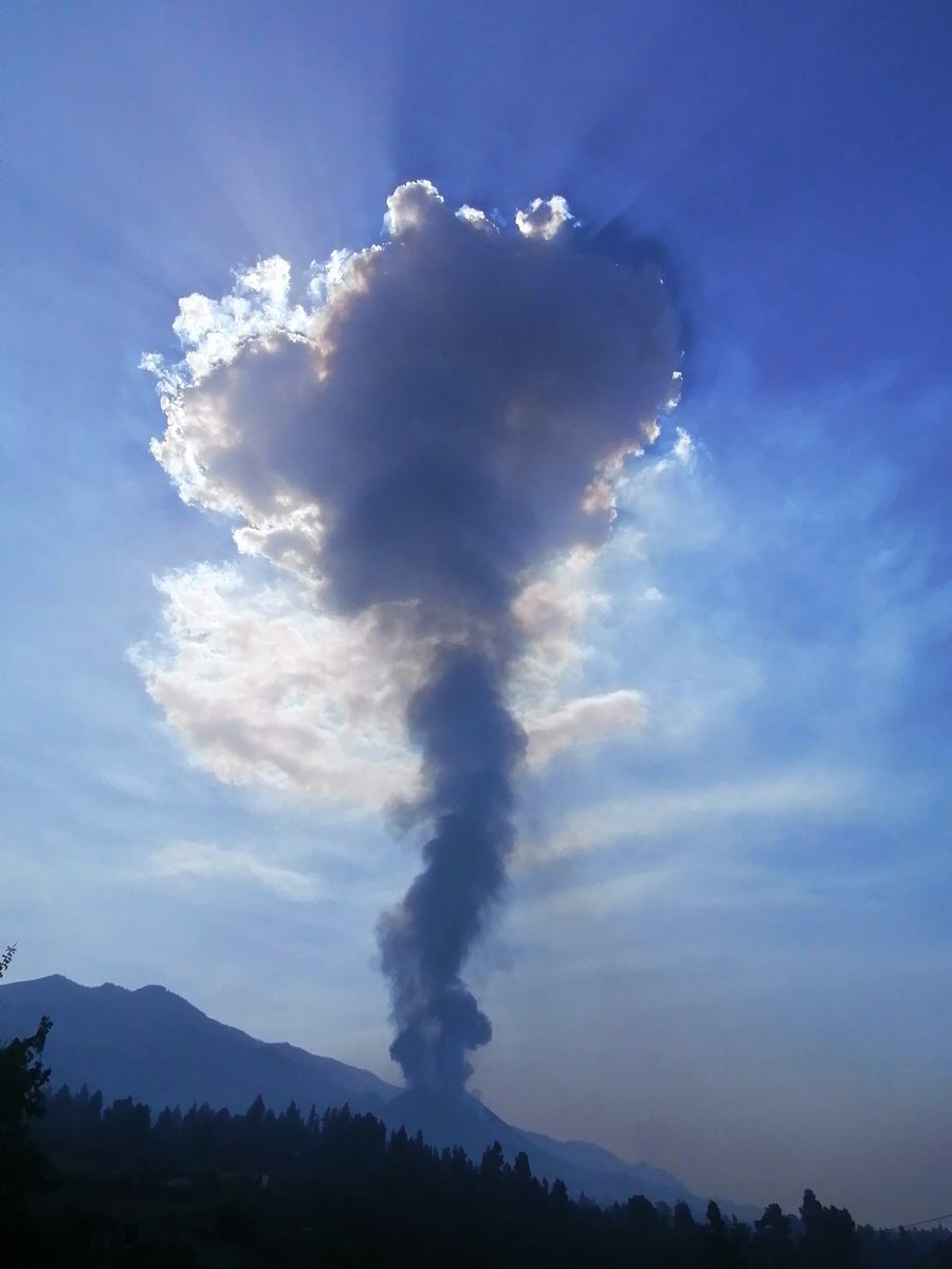 An erupting volcano, showing a tall grey ash plume extending into the blue sky.