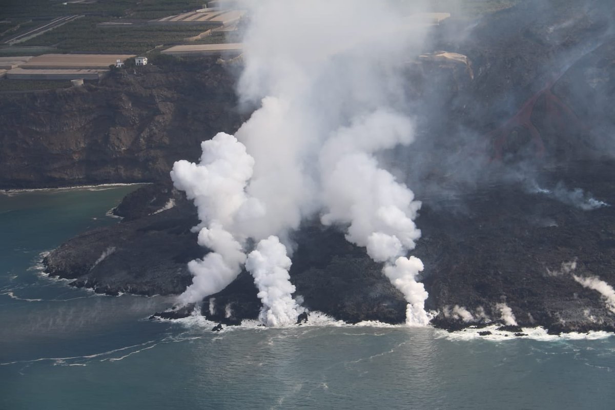 A flow of lava extending into the sea in the foreground, with two red lava flows feeding it. When the flow hits the sea, dense white clouds of vapour are seen.