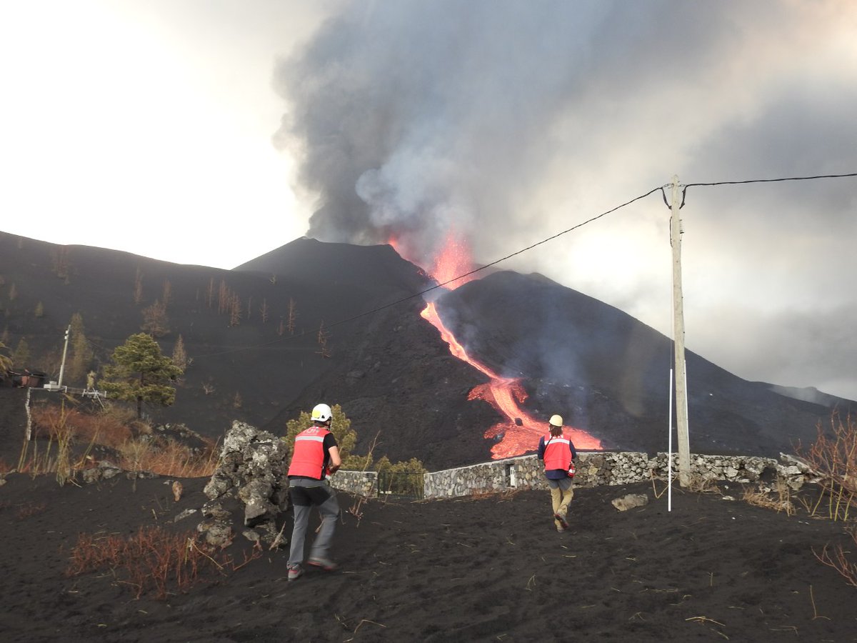 Scientists in high-vis look at the volcanic cone in the mid-ground. There is lots of ash rising from the top, and a 'waterfall' of orange lava flows are seen flowing down the flank of the cone.