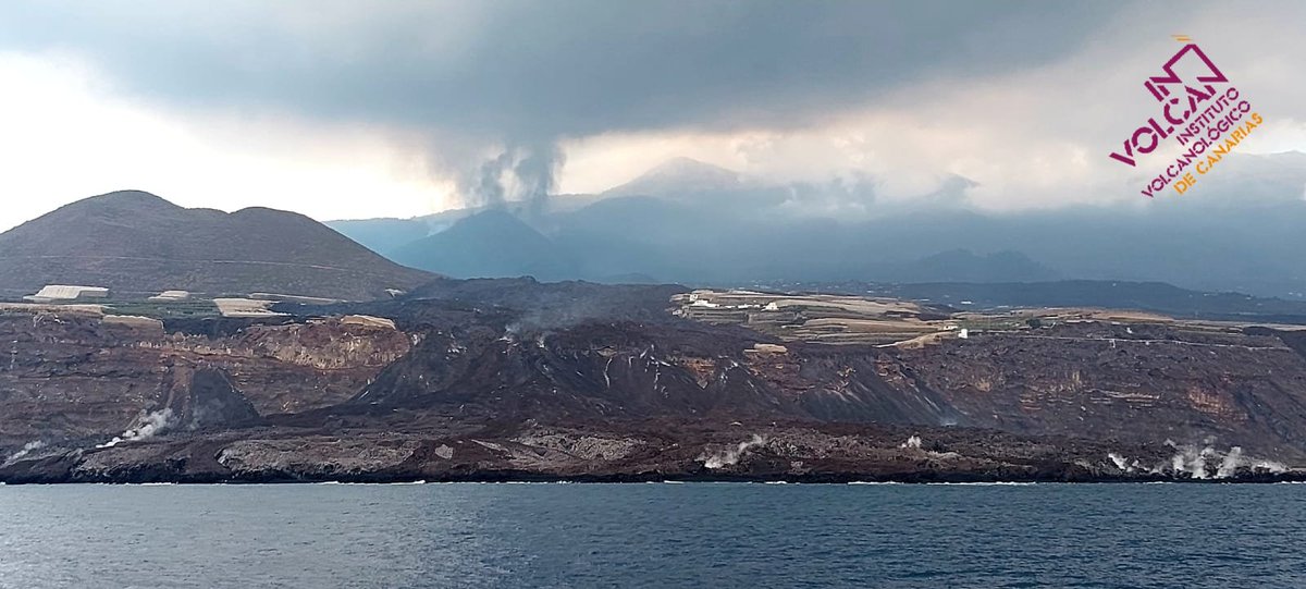 A picture from the sea looking towards the cliffs where lava has fallen down, with the lava flows and volcano seen in the background, with multiple ash plumes being seen