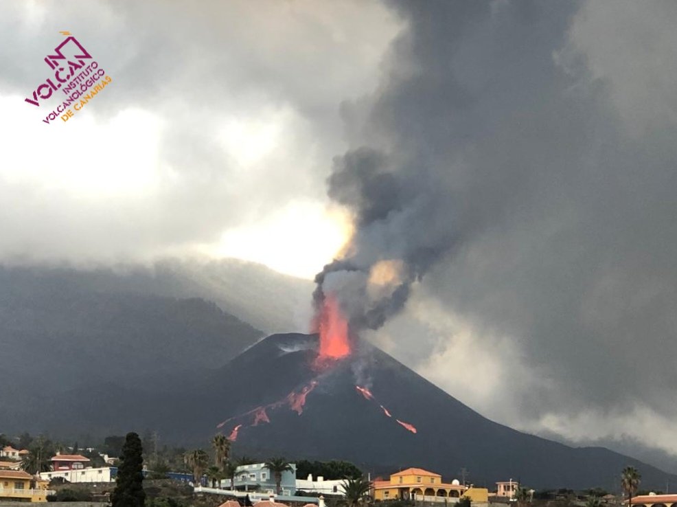 The main black cone of the volcano rises in the middle background, emitting an orange jet of lava and ash above that. Small streams of lava are seen flowing down the flanks of the cone. There are buildings in the foreground and lots of clouds around the volcano.