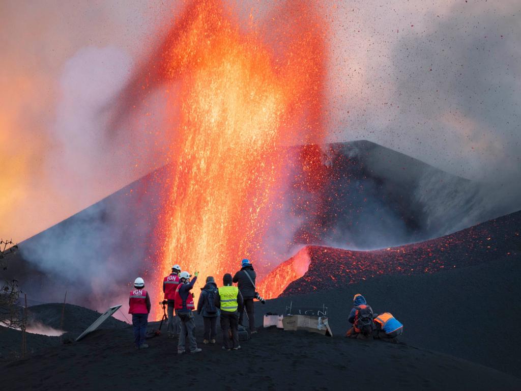 A group in high-visibility vests stand on grey ash in front of a large volcanic cone with an orange jet of lava coming out of it. There are various wires and pieces of equipment on the floor which are being attended to.