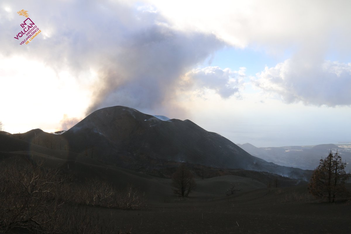The large dark grey cone of the volcano is seen with some steam rising from the top but no lava or ash. In the background the black lava flow field can be seen extending towards the sea