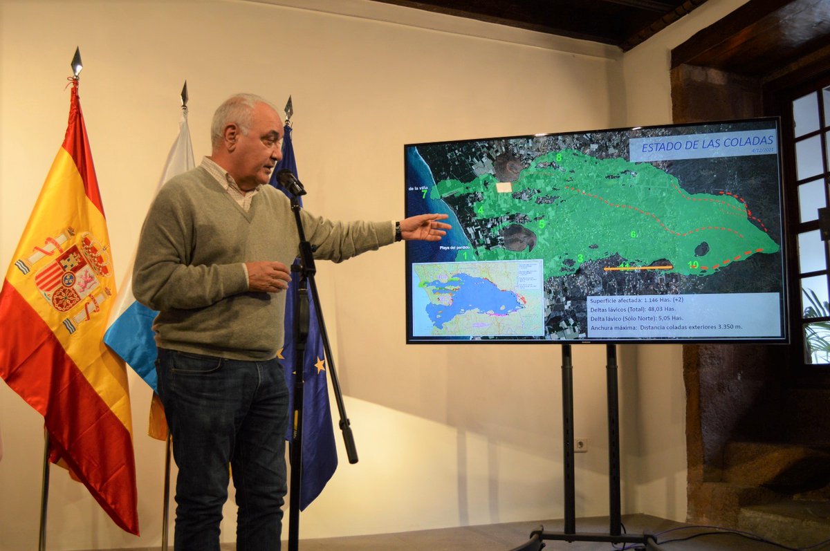 A man in front of three flags (Spain, Canary Islands and European Union) gestures to a TV screen where the latest lava flow extents are shown