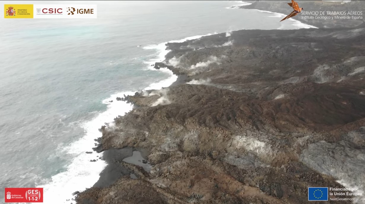 A smoking, rubblely mass of brown and black lava stick out into the sea. Breaking waves are seen at the edge of the lava flows, where erosion has formed beaches